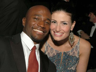Taye Diggs and Idina Menzel are touching their cheeks as they are smiling at the camera.